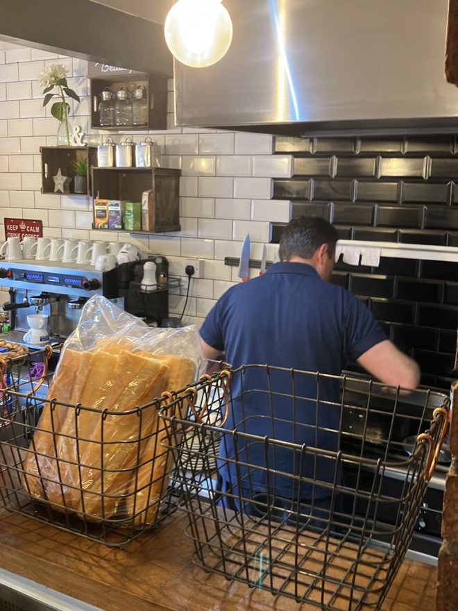 A staff member working at the white-tiled kitchen pass, with a wire basket of wrapped bread in the foreground.